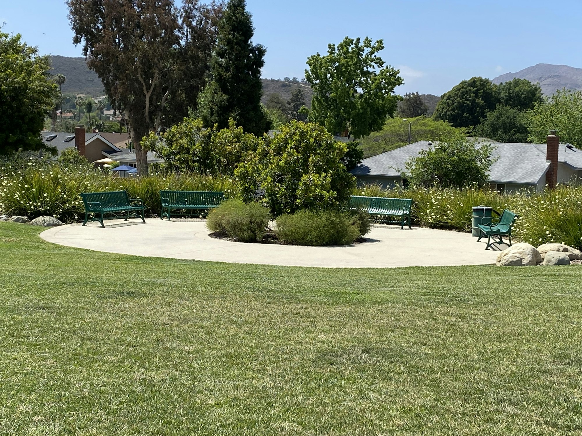 A park with a circular paved area, green benches, trees, and houses in the background.