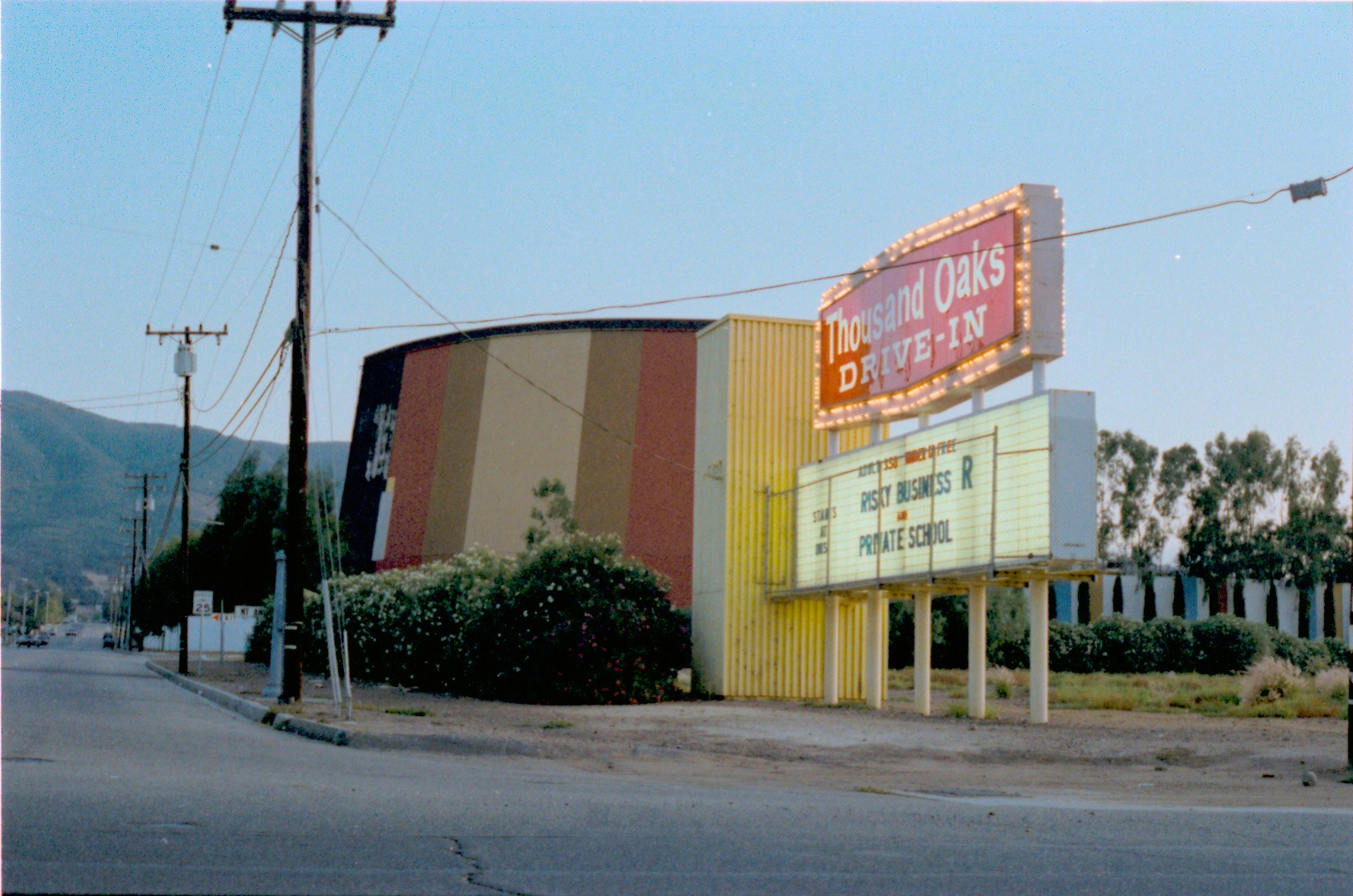 Thousand Oaks Drive-In sign, marquee reads "Risky Business," large screen, power lines, and road with trees and mountains in the background.