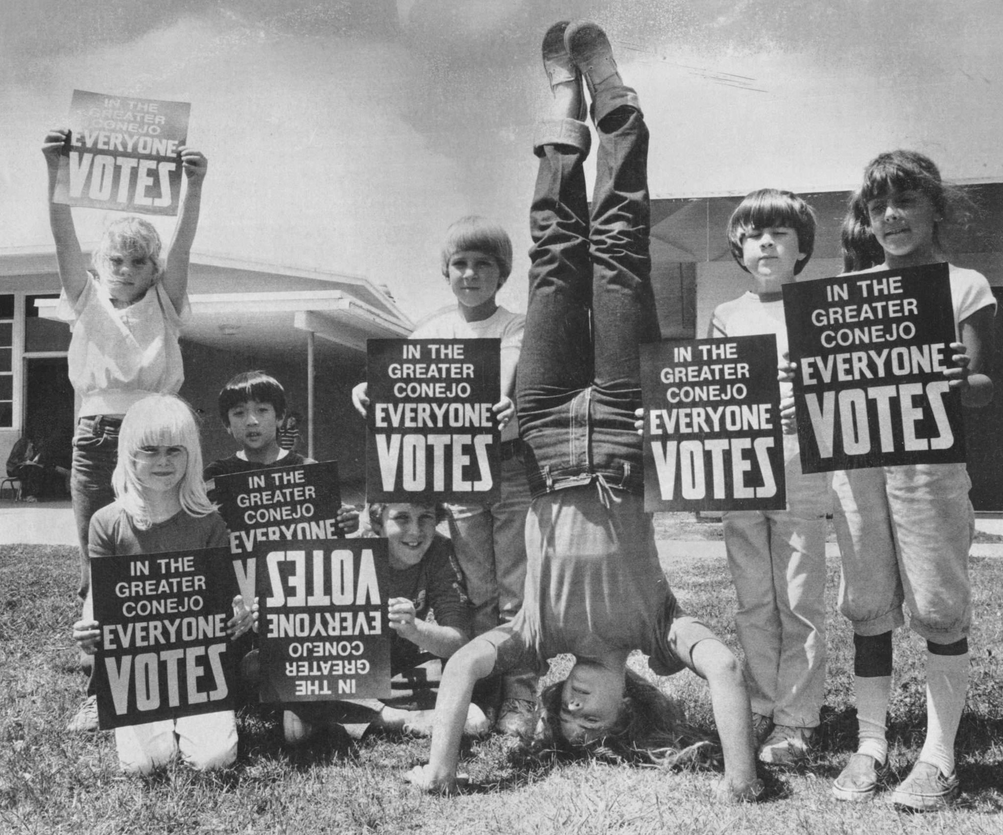 Children holding "In the Greater Conejo Everyone Votes" signs, with one child doing a handstand in the center.