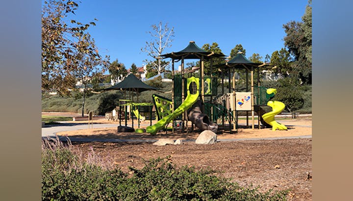 A playground with slides, climbing structures, and a sandy play area surrounded by trees and grass under a clear blue sky.
