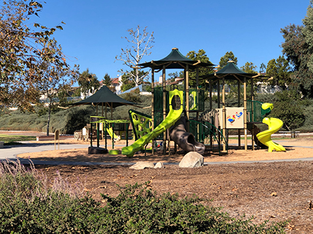 A playground with slides, climbing structures, and a sandy play area surrounded by trees and grass under a clear blue sky.