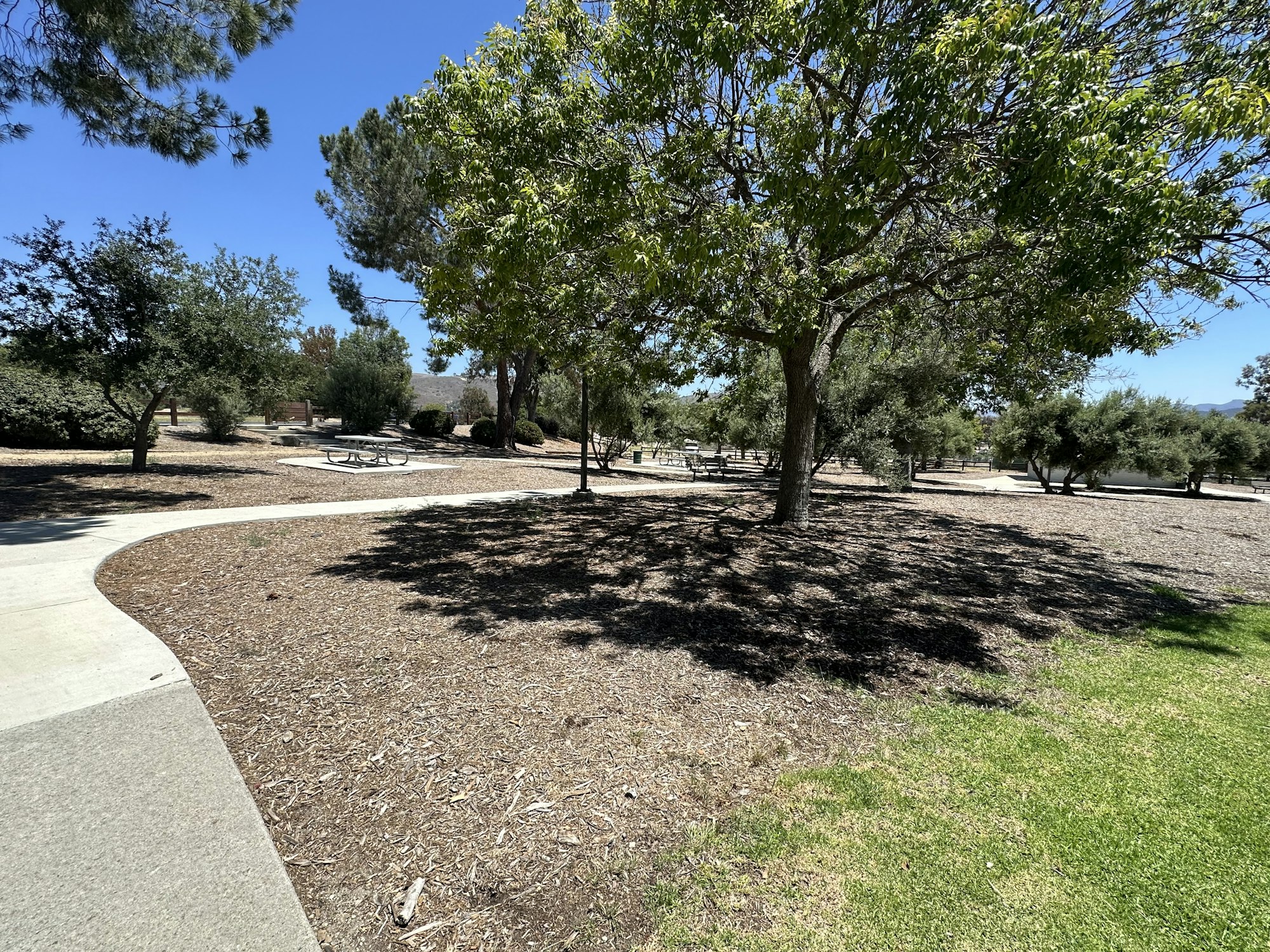 The image shows a park with walking paths, trees, picnic tables, and sunny blue skies, creating a serene outdoor setting.