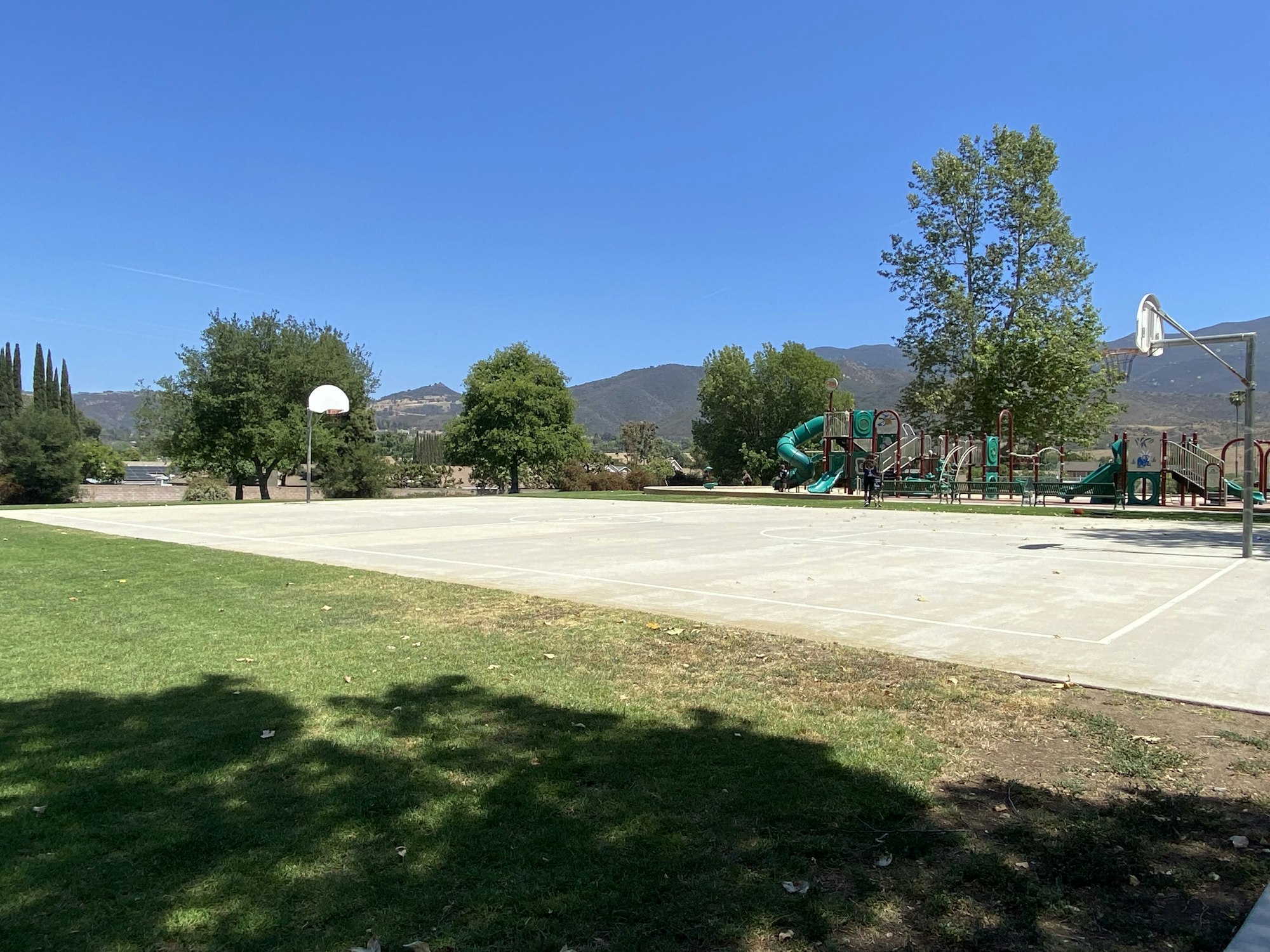 A park with a basketball court, a playground, trees, and mountains in the background under a clear blue sky.