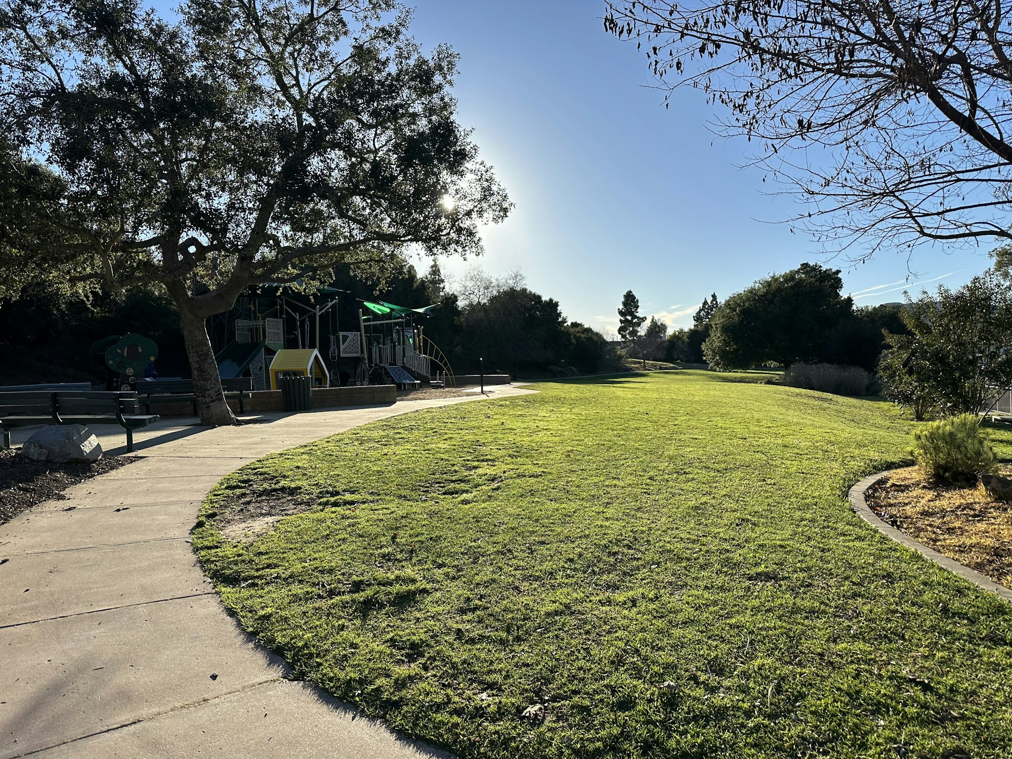 Park with a playground, trees, grass, and a curved path under a clear blue sky.