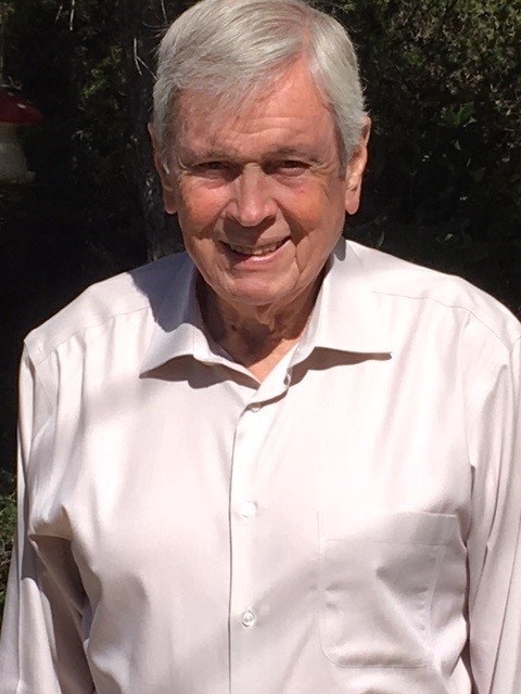 An older man in a light shirt smiling outdoors with trees in the background.