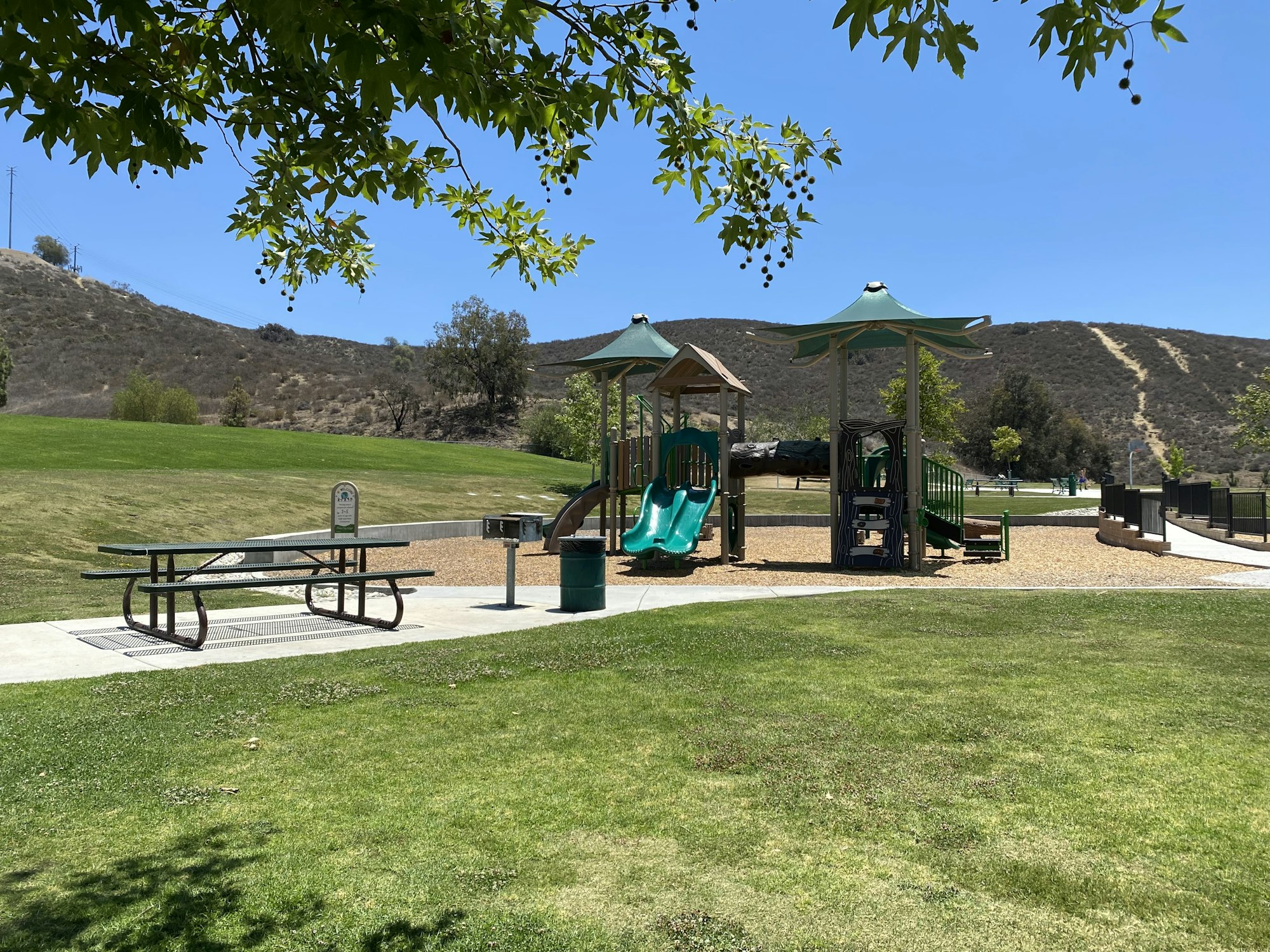 Playground with green slides, picnic table, and grill in a park with hills and trees in the background.