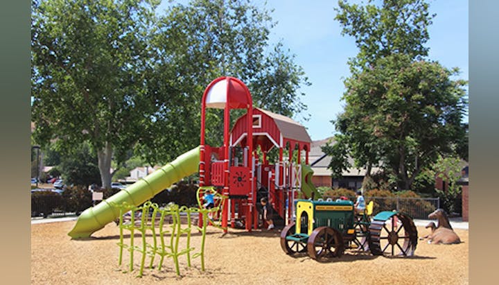 A colorful playground with slides, climbing structures, a tractor-themed play area, and trees in the background.
