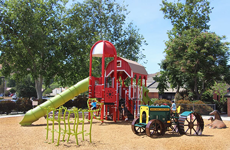 A colorful playground with slides, climbing structures, a tractor-themed play area, and trees in the background.