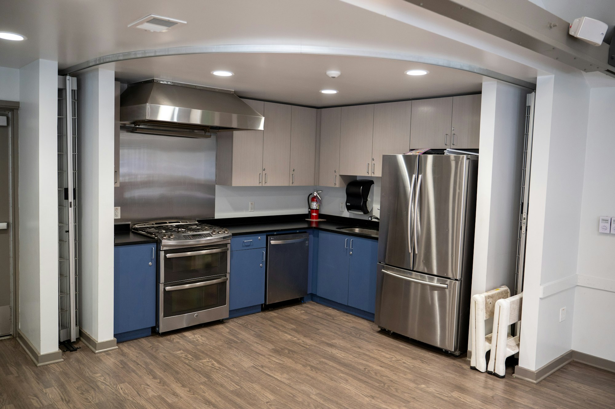 Modern kitchen with stainless steel appliances, blue cabinets, and a fire extinguisher on a wood floor.