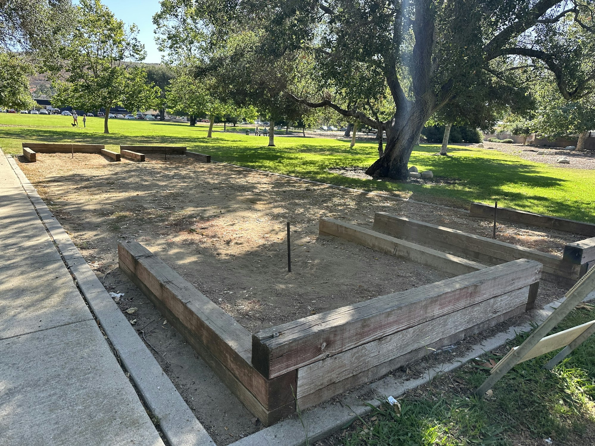 A cleared area with wooden garden beds in a park, surrounded by grass and trees, and a sidewalk nearby.