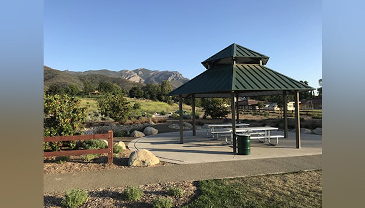 A gazebo with picnic tables, surrounded by greenery and mountains under a clear blue sky.