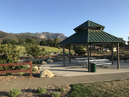 A gazebo with picnic tables, surrounded by greenery and mountains under a clear blue sky.