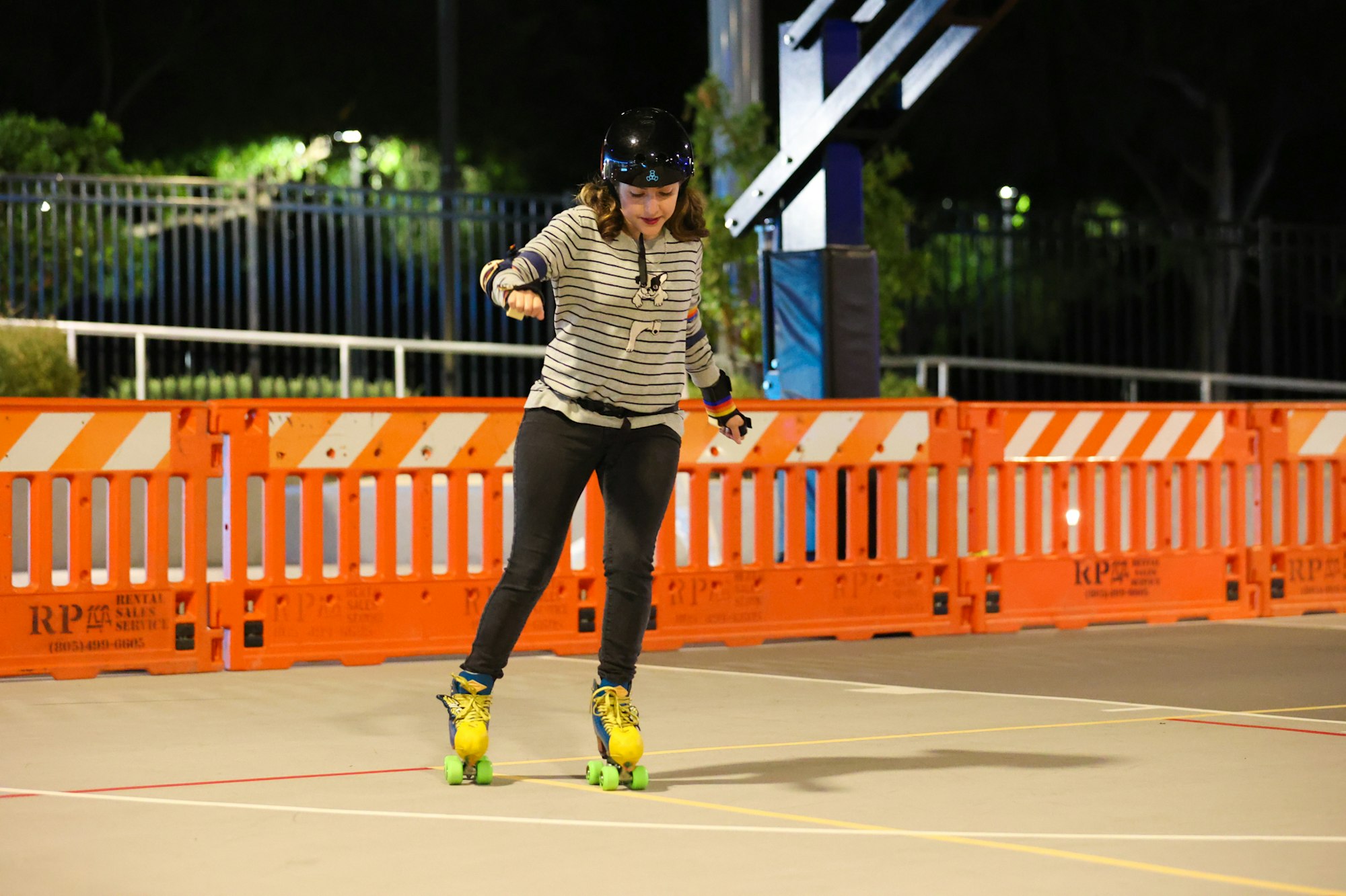 A girl roller skating at night, wearing a helmet and protective gear, focused on balancing in front of orange barricades.