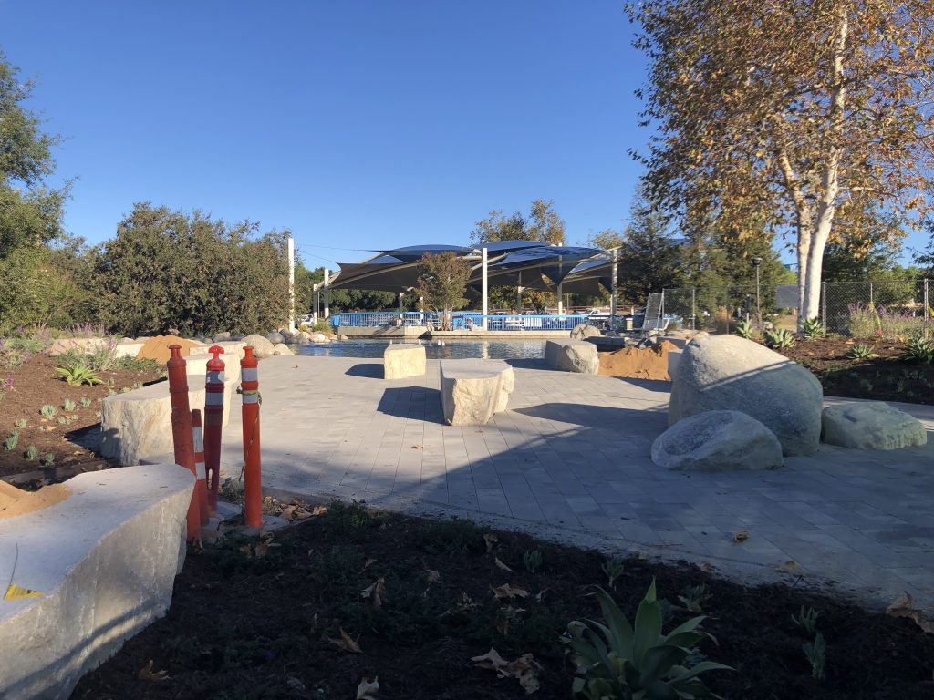 Outdoor space with paved path, large rocks, greenery, red posts, and a shaded area in the background.