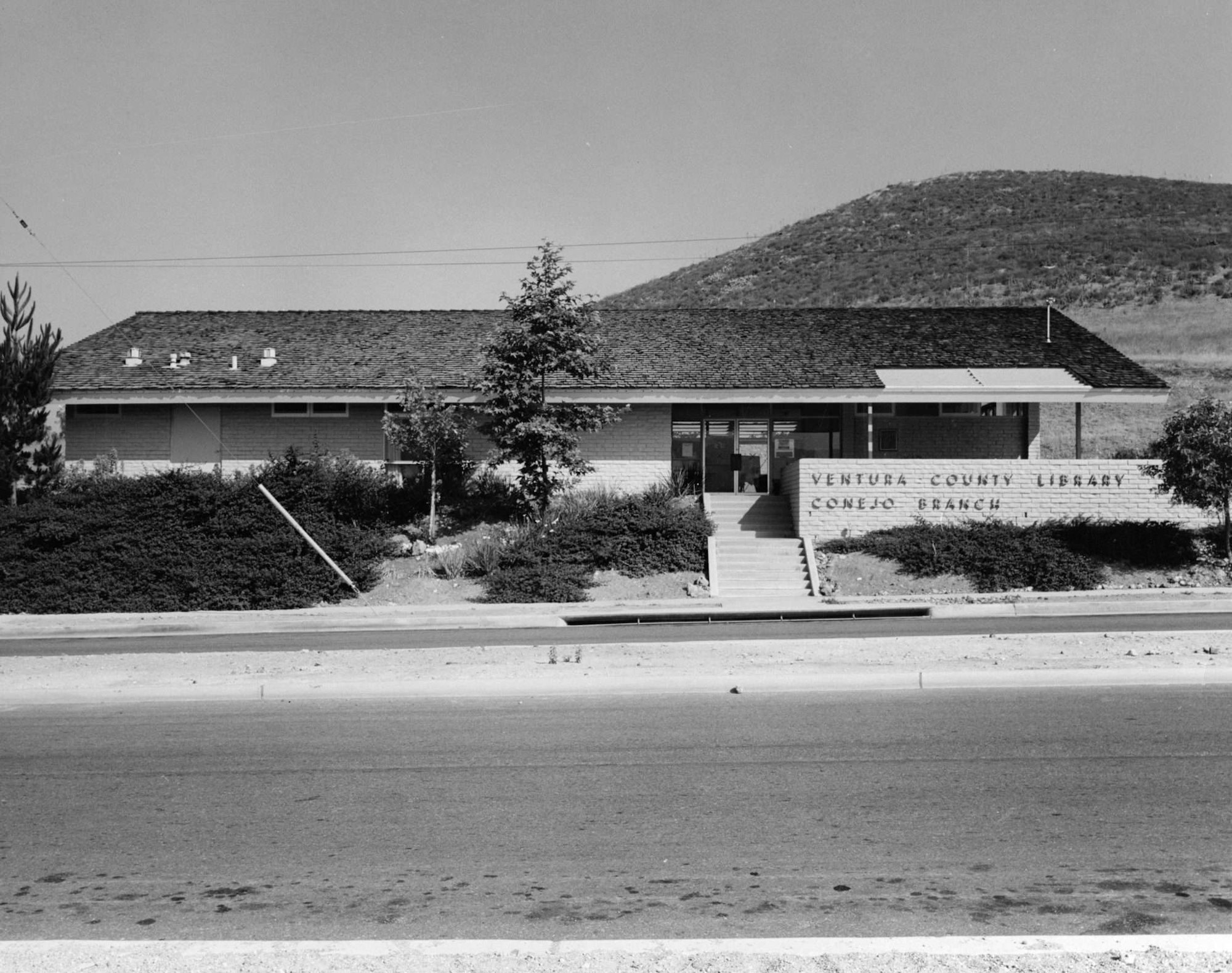 A black-and-white photo of Ventura County Library, Conejo Branch, with a tree and hills in the background.