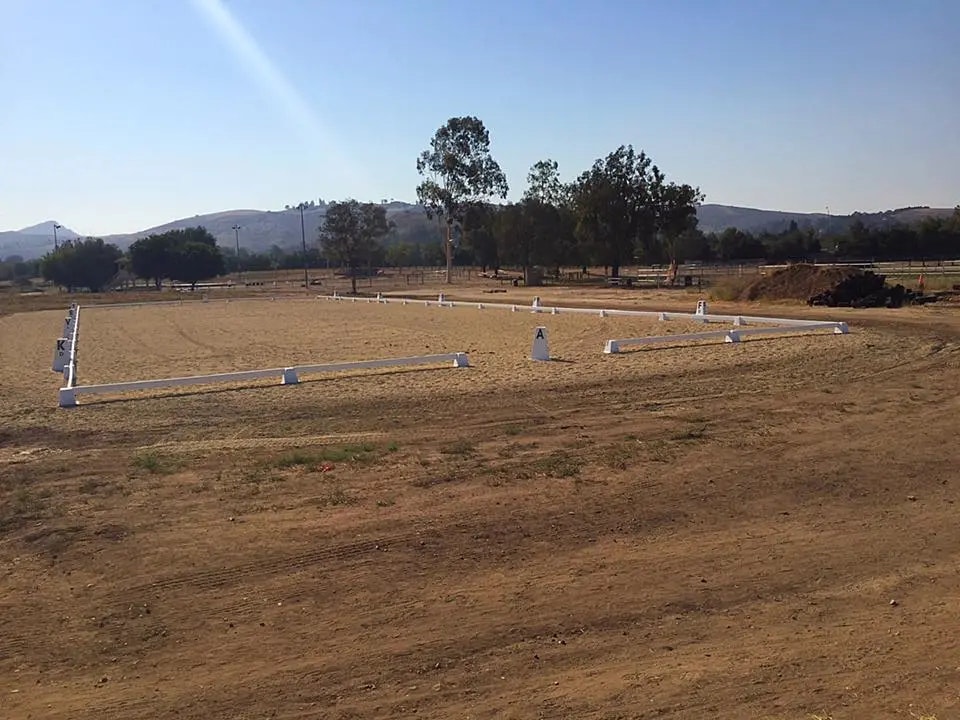 An empty dressage arena with letter markers, surrounded by trees and hills under a clear sky.