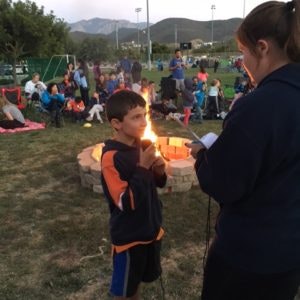 A child holds a marshmallow on a stick by a fire pit, while a person supervises, with a crowd in the background and mountains visible.