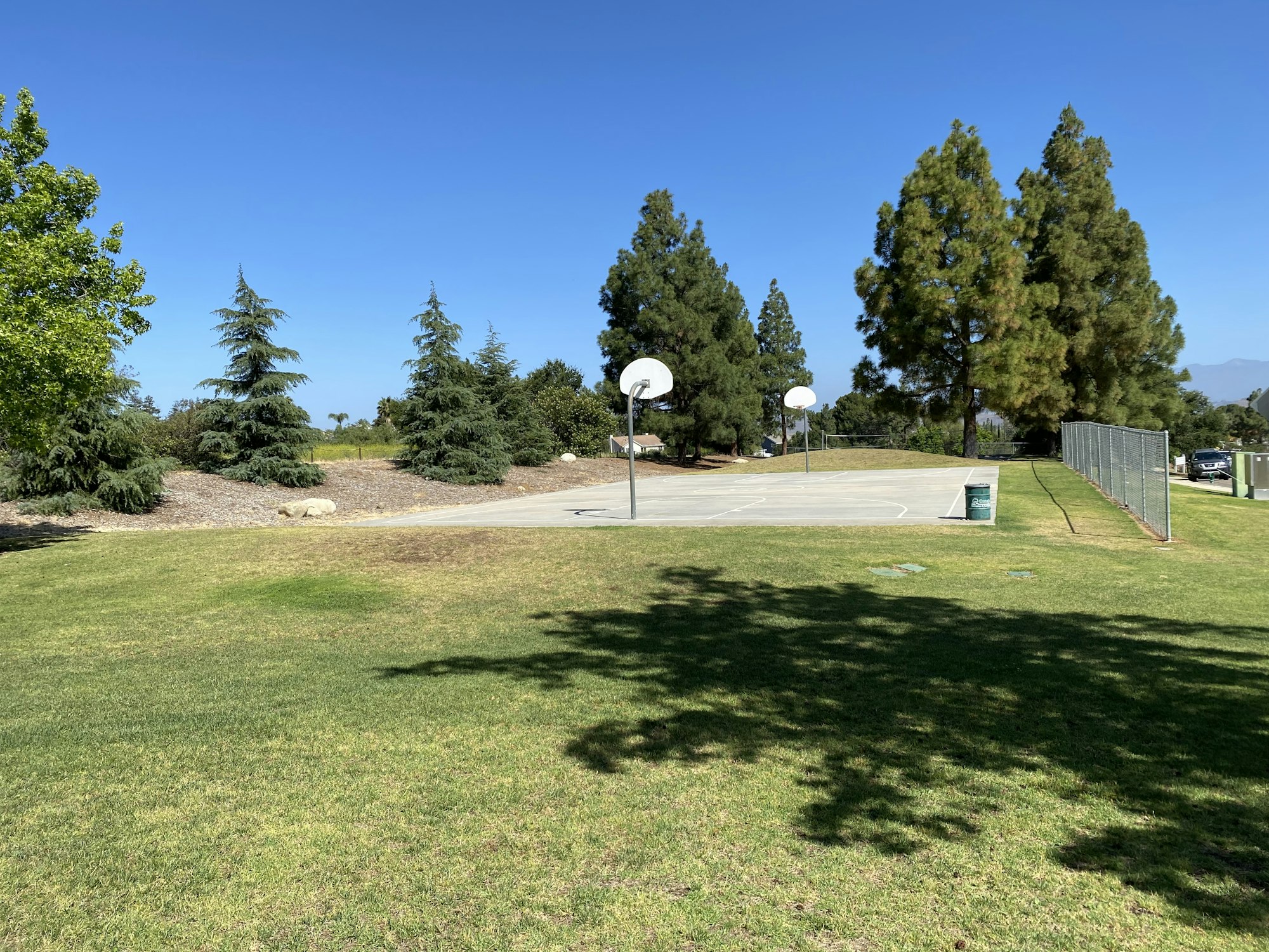 Basketball court with hoops, surrounded by grass and trees under a clear blue sky.