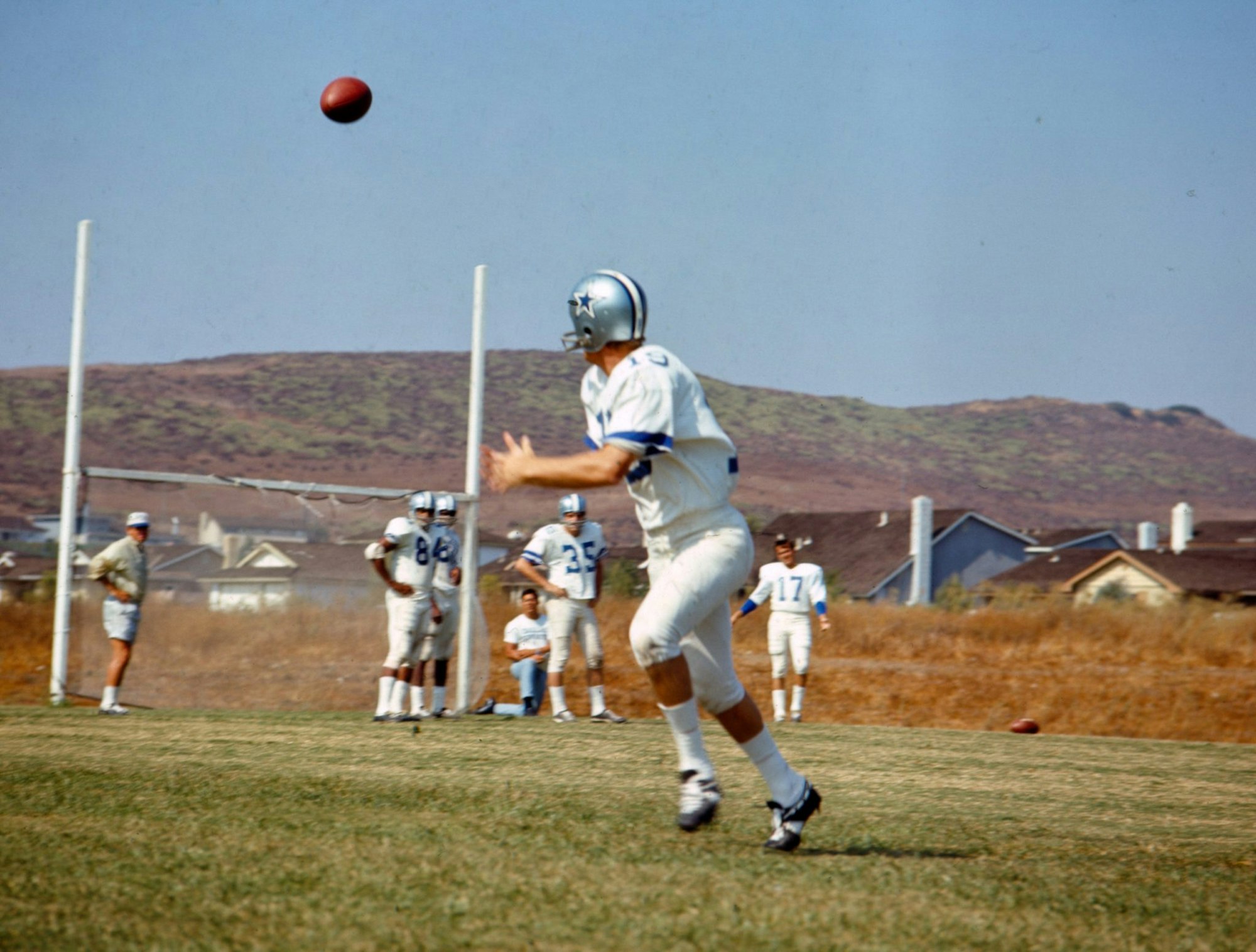 Football player in white uniform and helmet catching a ball during practice on a field with hills in the background.
