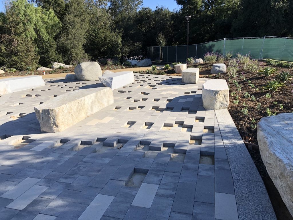 Stone benches and large rocks on a geometric paved area, surrounded by trees and a green fence.