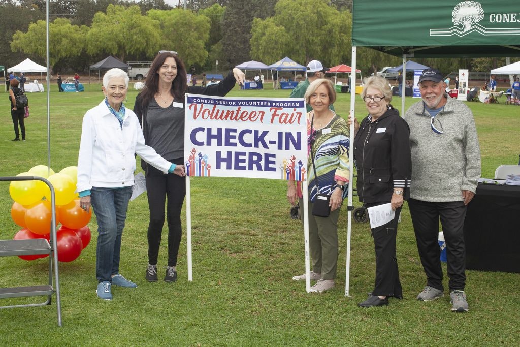 People standing by a "Volunteer Fair Check-In Here" sign at an outdoor event with tents and balloons.