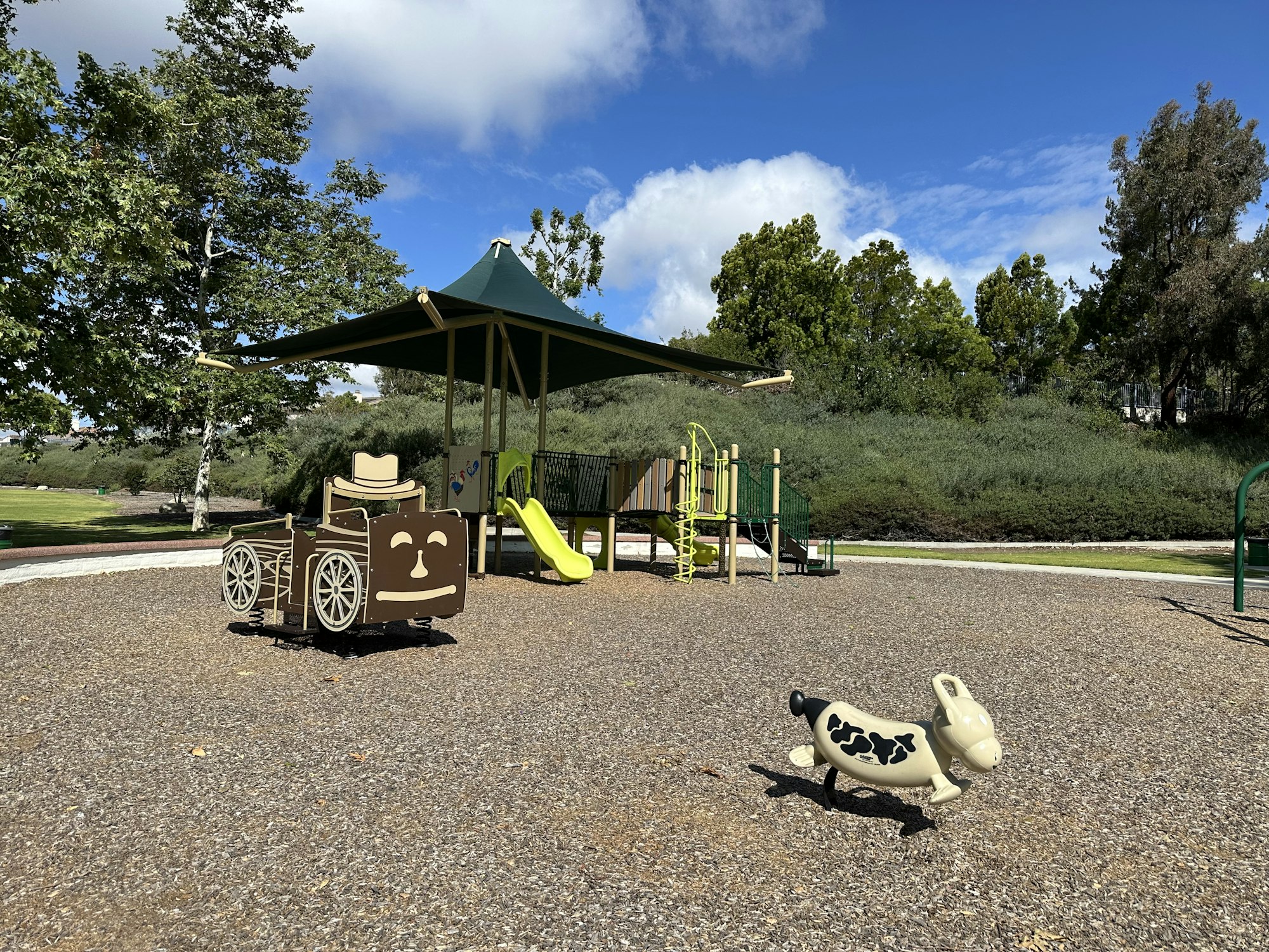 The image shows a playground with a smiling wheelchair and a playful cow-shaped spring rider under a covered area.