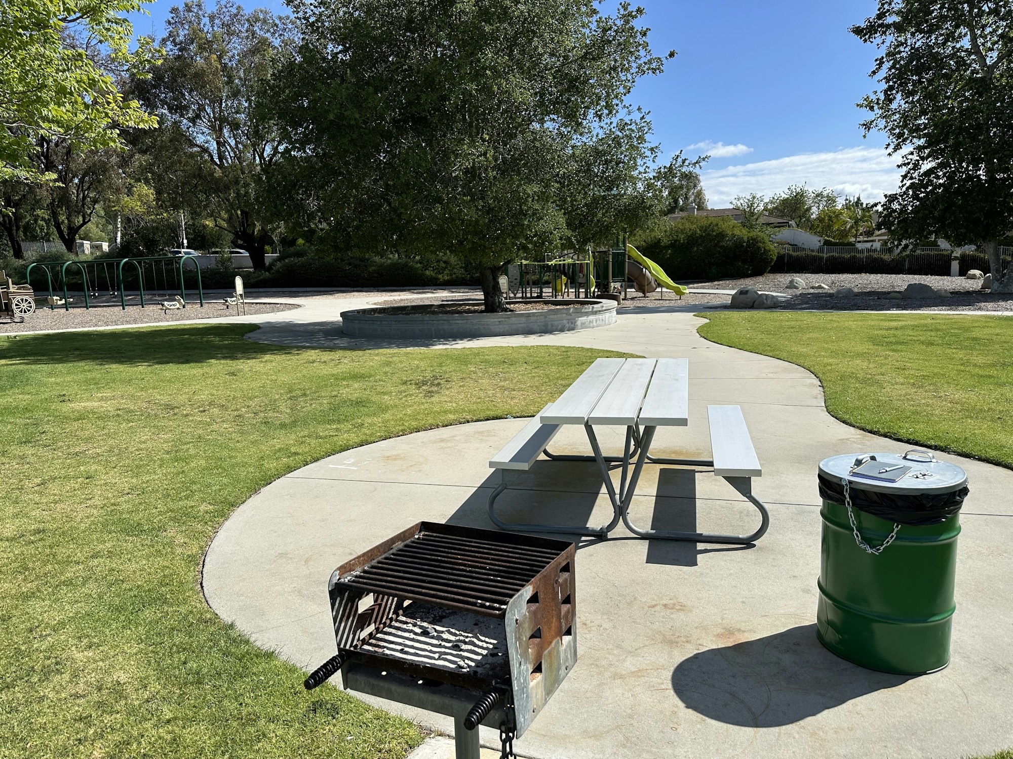 A park scene featuring a picnic table, grill, trash can, playground equipment, and lush green grass under a blue sky.