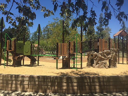 Playground with slides, climbing structures, and wood chips, surrounded by trees under a clear blue sky.
