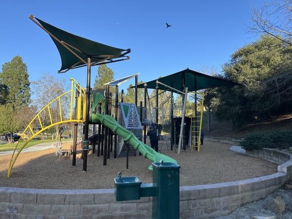 A playground with slides, climbing structures, and shade canopies set in a park with trees and clear blue sky.