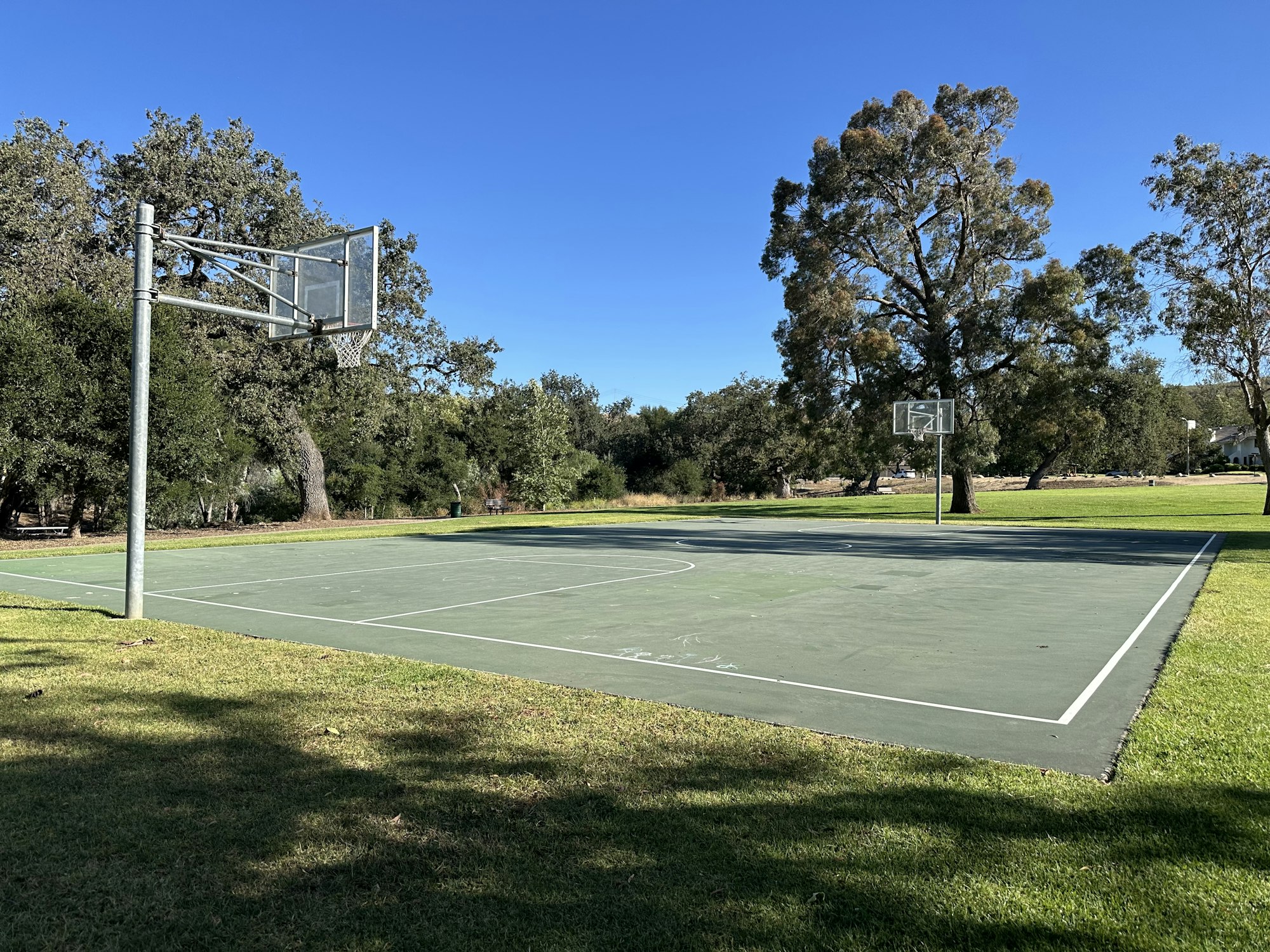 A vacant outdoor basketball court surrounded by trees under a clear blue sky.