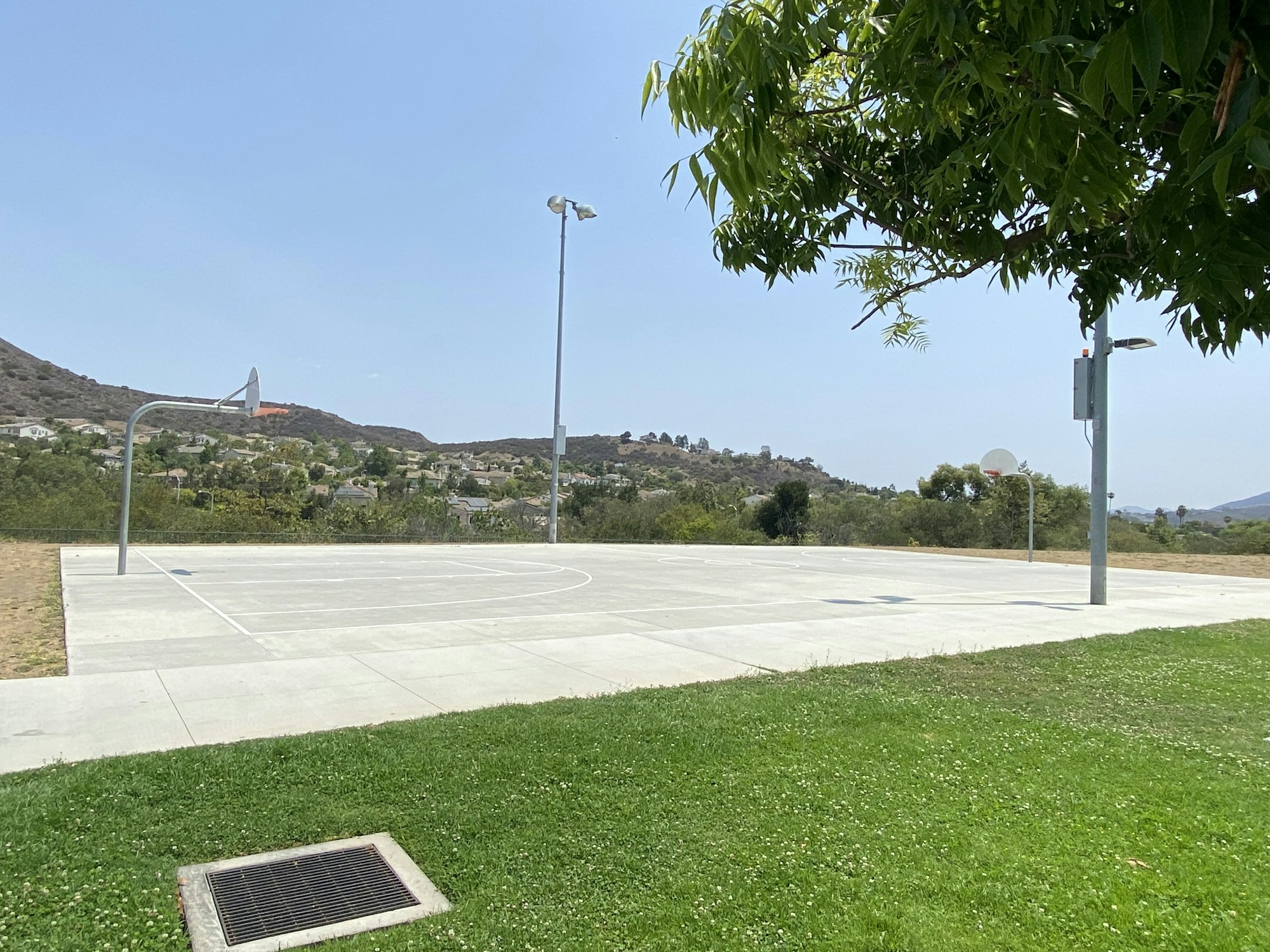 Outdoor basketball court in a park setting, surrounded by grass and hills, with two hoops and light poles.