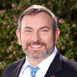 A smiling man in a suit and tie with a blurred floral background.
