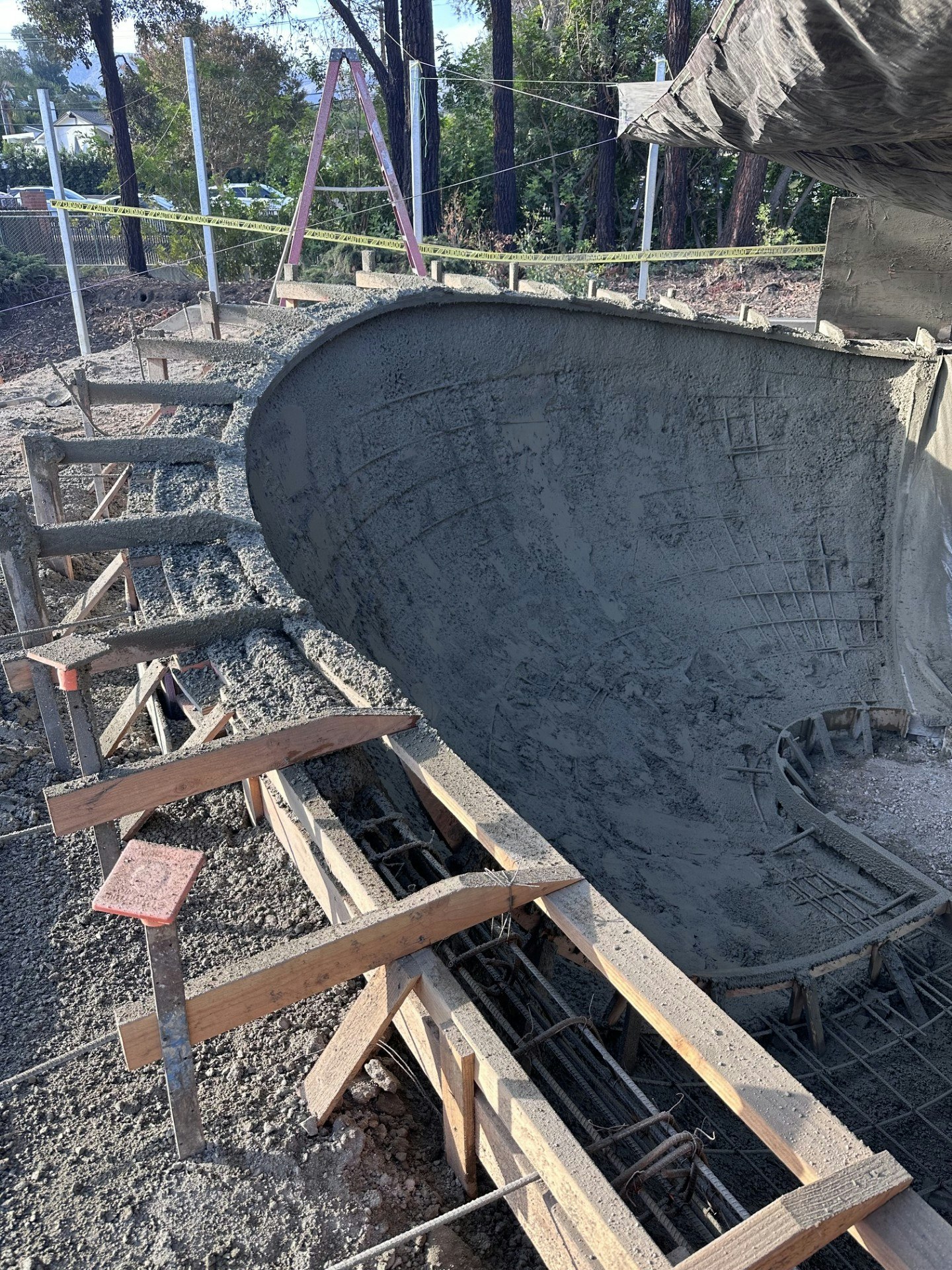 A construction site with a freshly poured concrete structure, featuring wooden frames and a curved shape, surrounded by trees.
