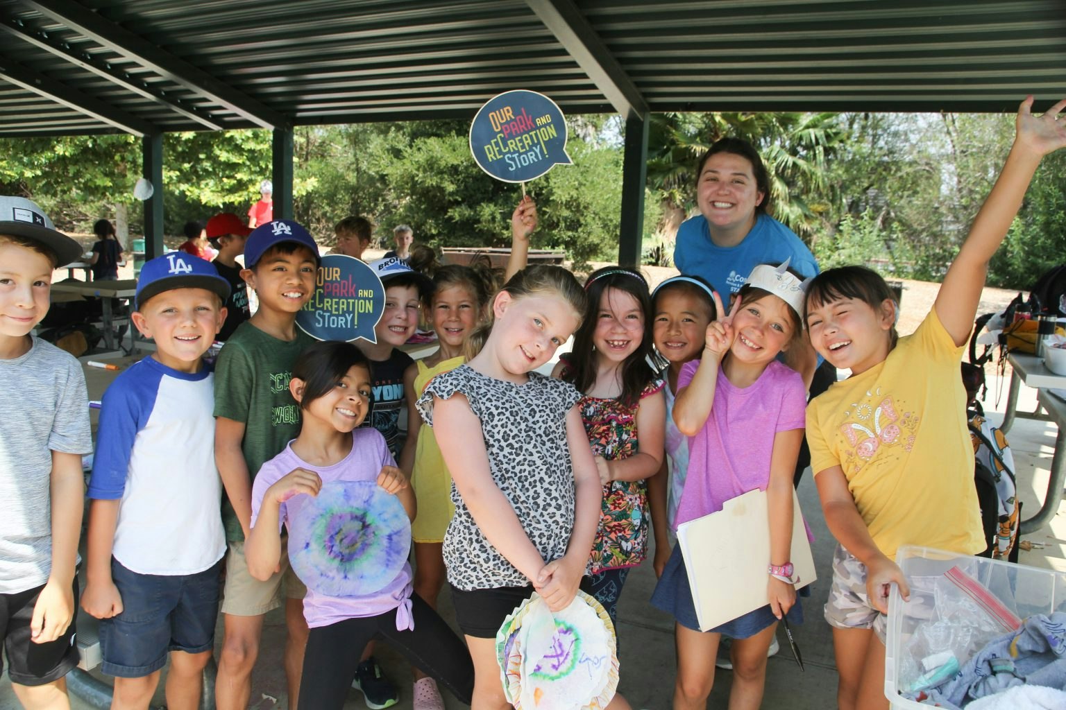 A group of joyful kids and a woman posing with "Our Park and Recreation Story" signs under a pavilion.