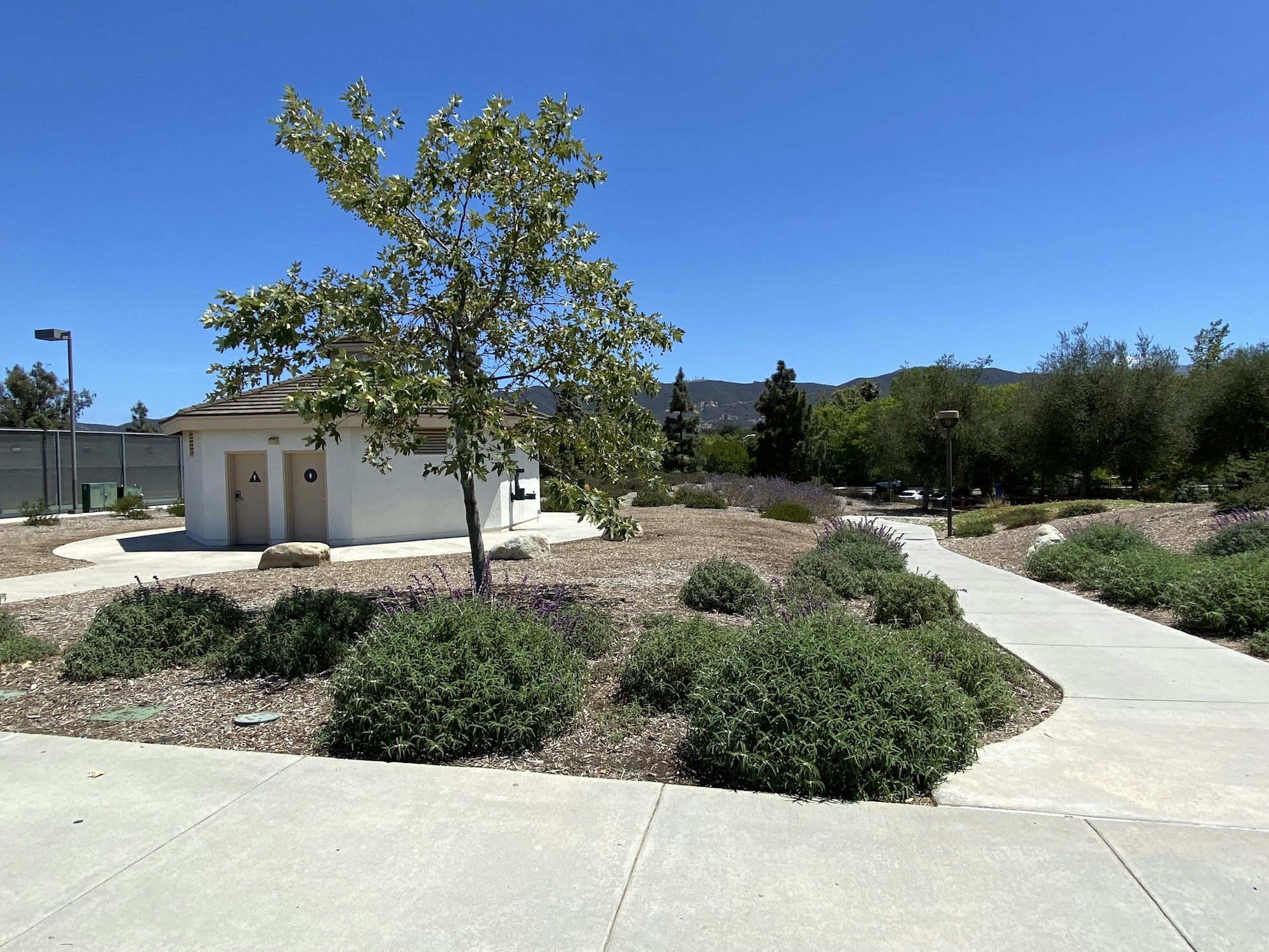 A park path leads to a restroom, surrounded by shrubs, trees, and open sky.