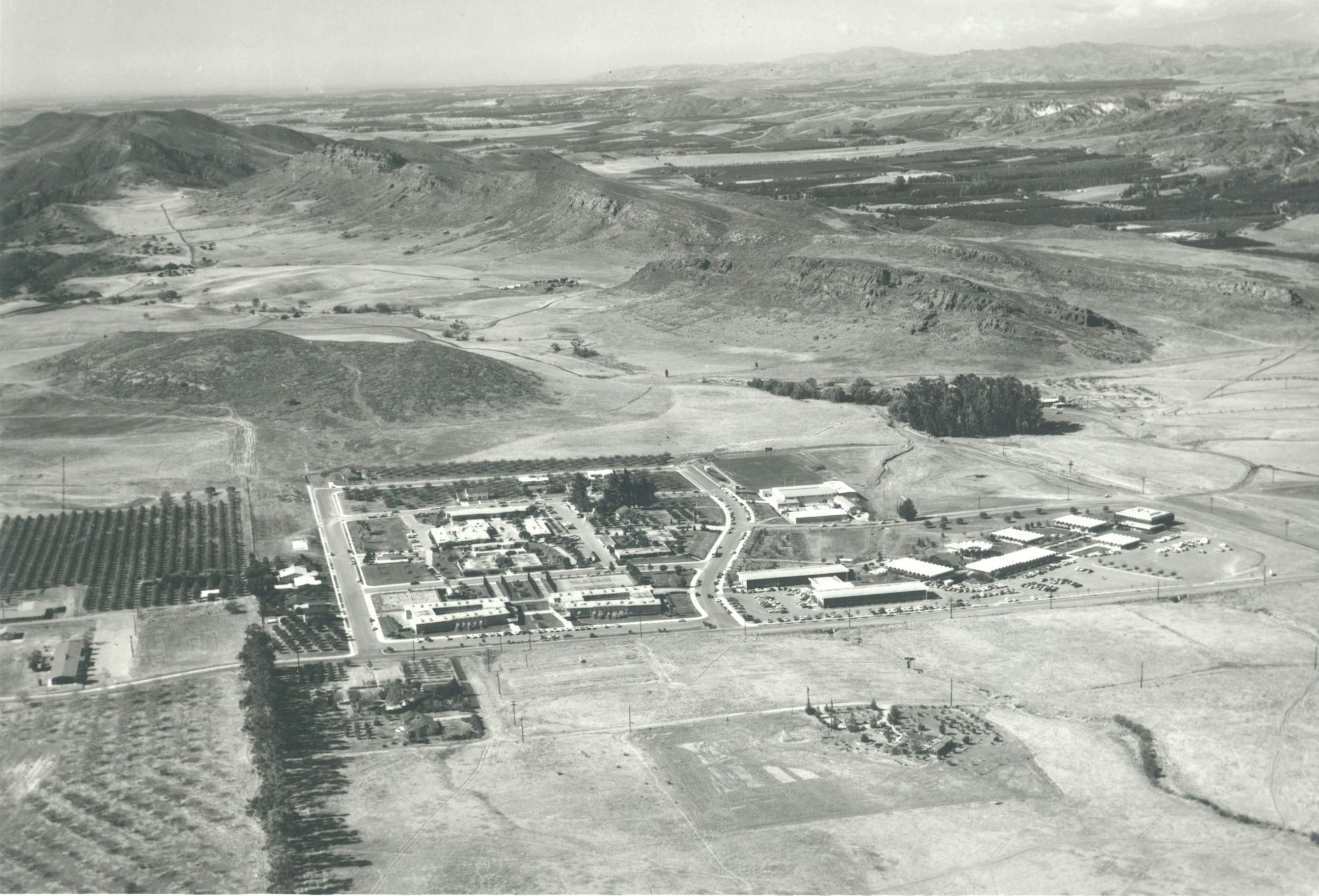 Aerial view of a small town or campus with buildings and parking, surrounded by hills and vast open fields.