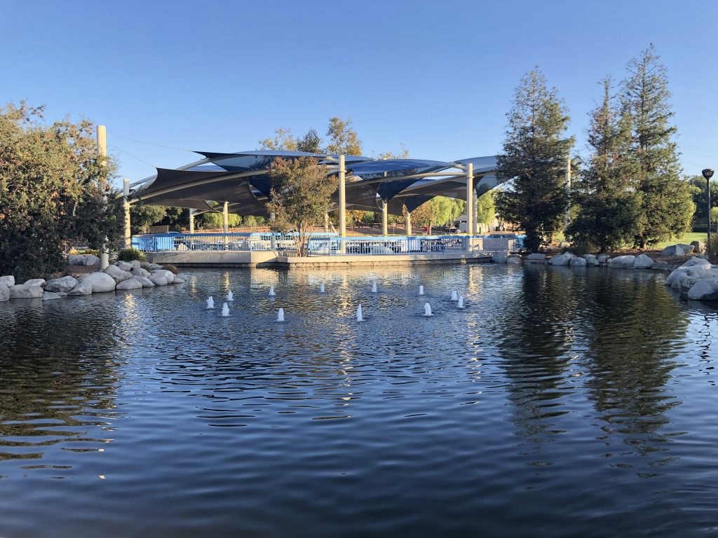 A pond with small fountains, surrounded by trees and rocks. There's a structure with shaded canopies in the background.