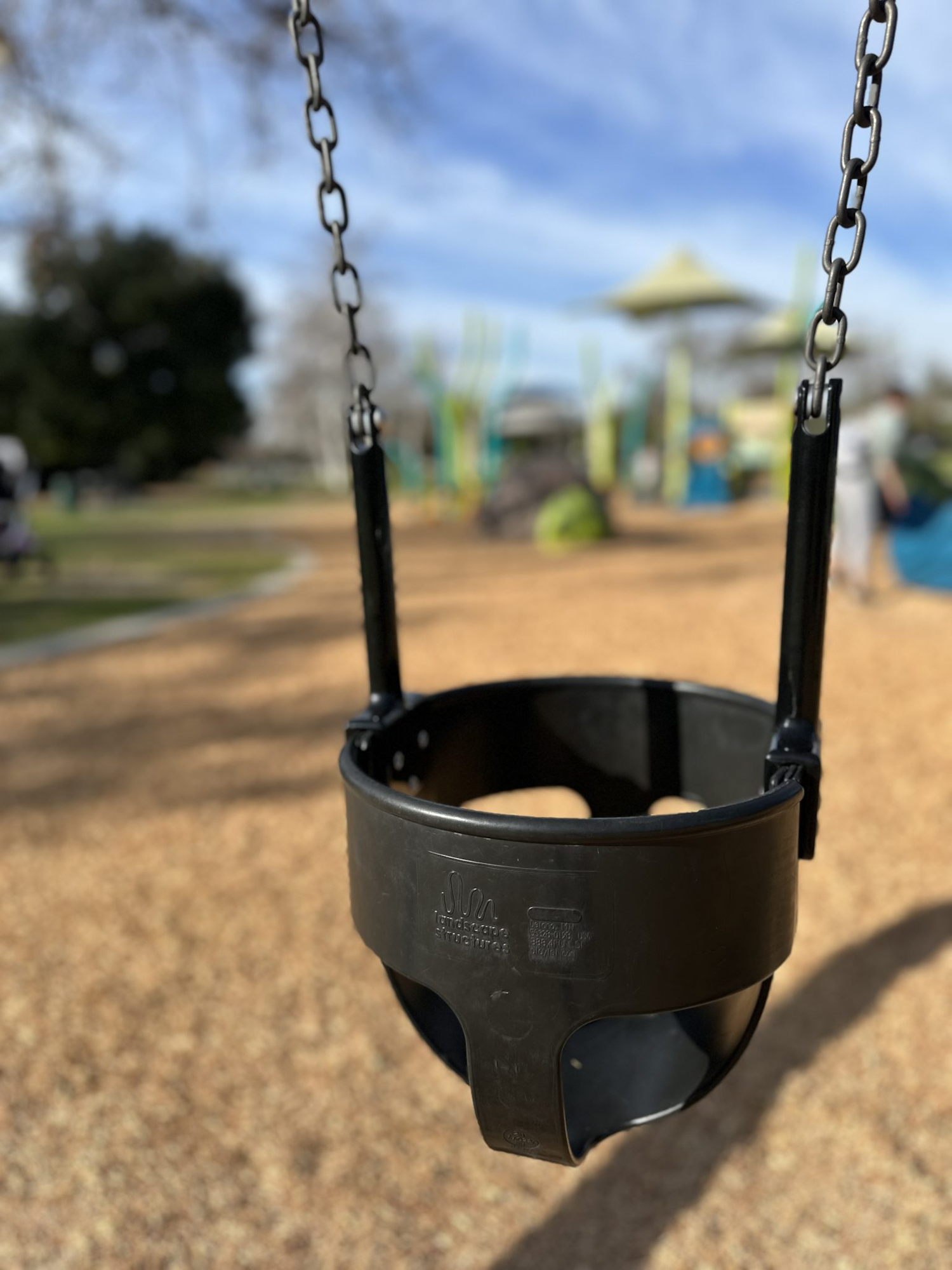A black toddler swing at a playground, with chains, blurred background of park and trees.