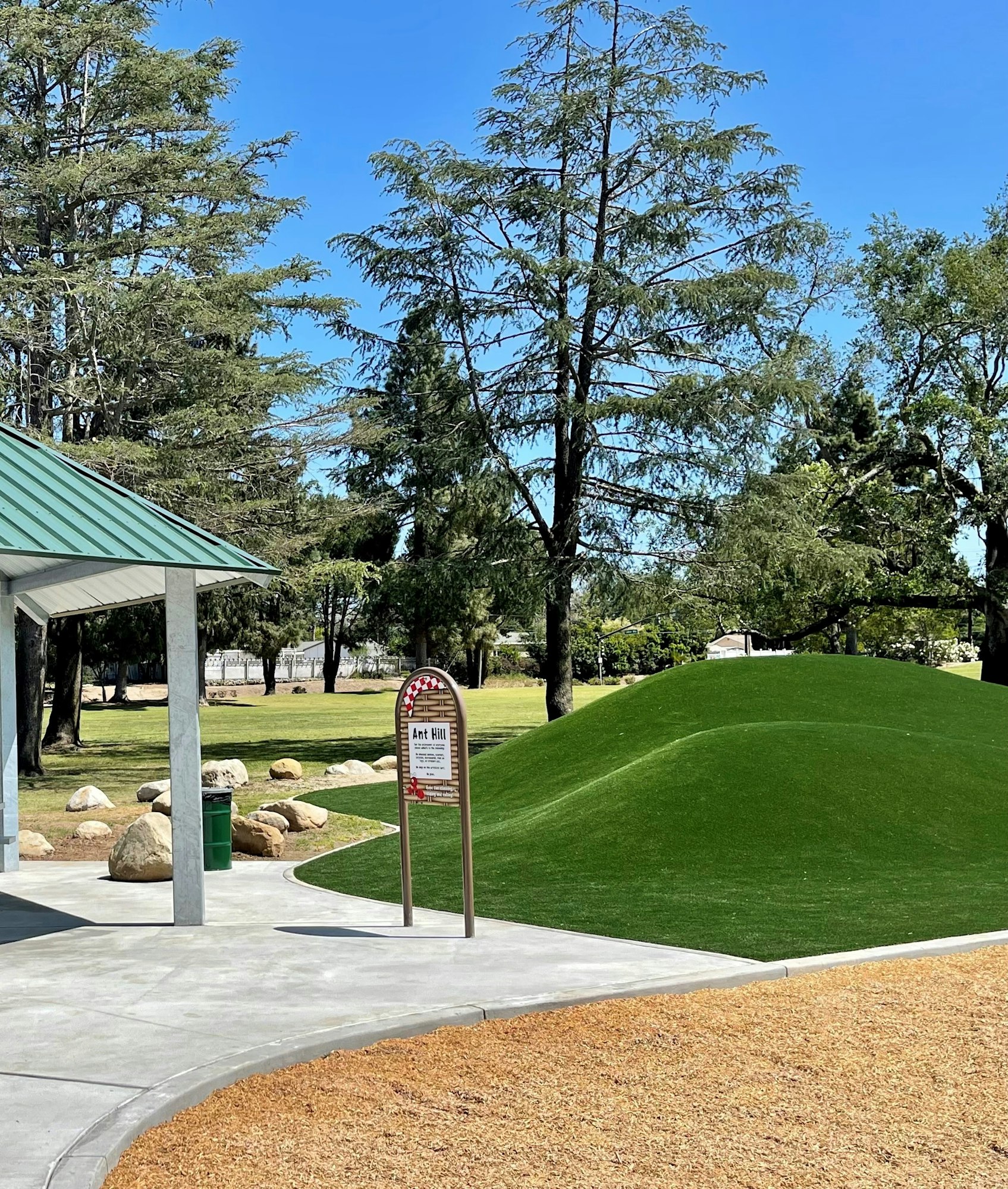 Park with pavilion, green artificial hill, pathway, trees, and a sign labeled "Ant Hill."