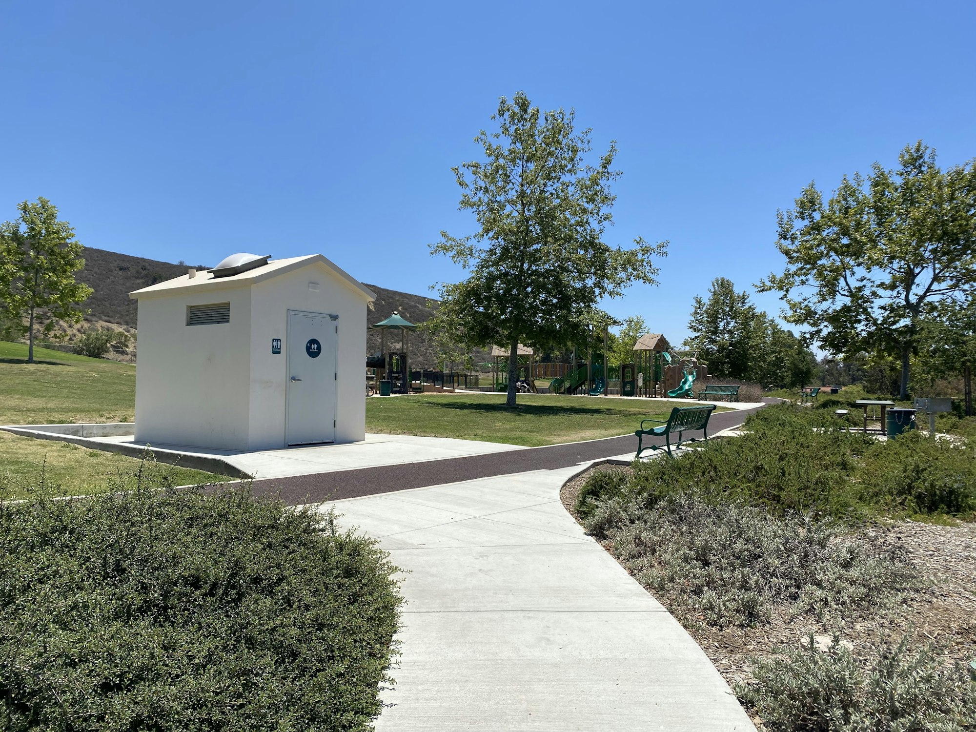 A restroom building, path, benches, playground, and trees in a park.