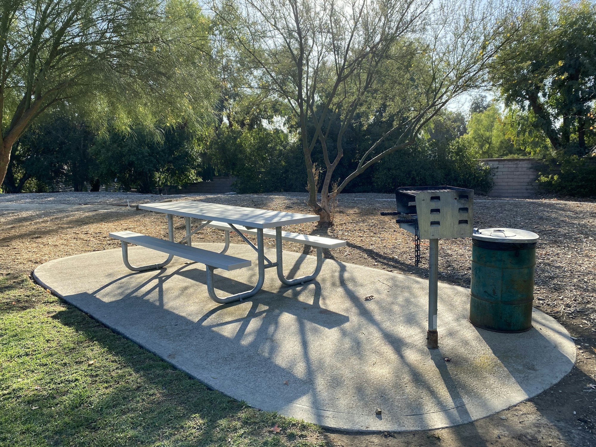 Picnic area with a bench, grill, and trash can under trees in a park.