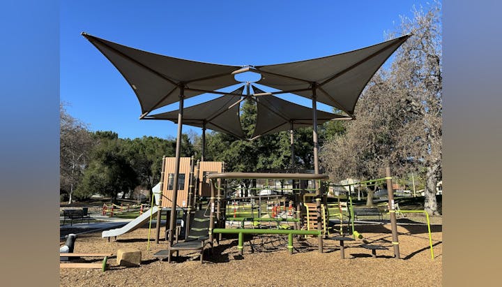Children's playground with slides, climbing structures, and large shade sails, set in a park.
