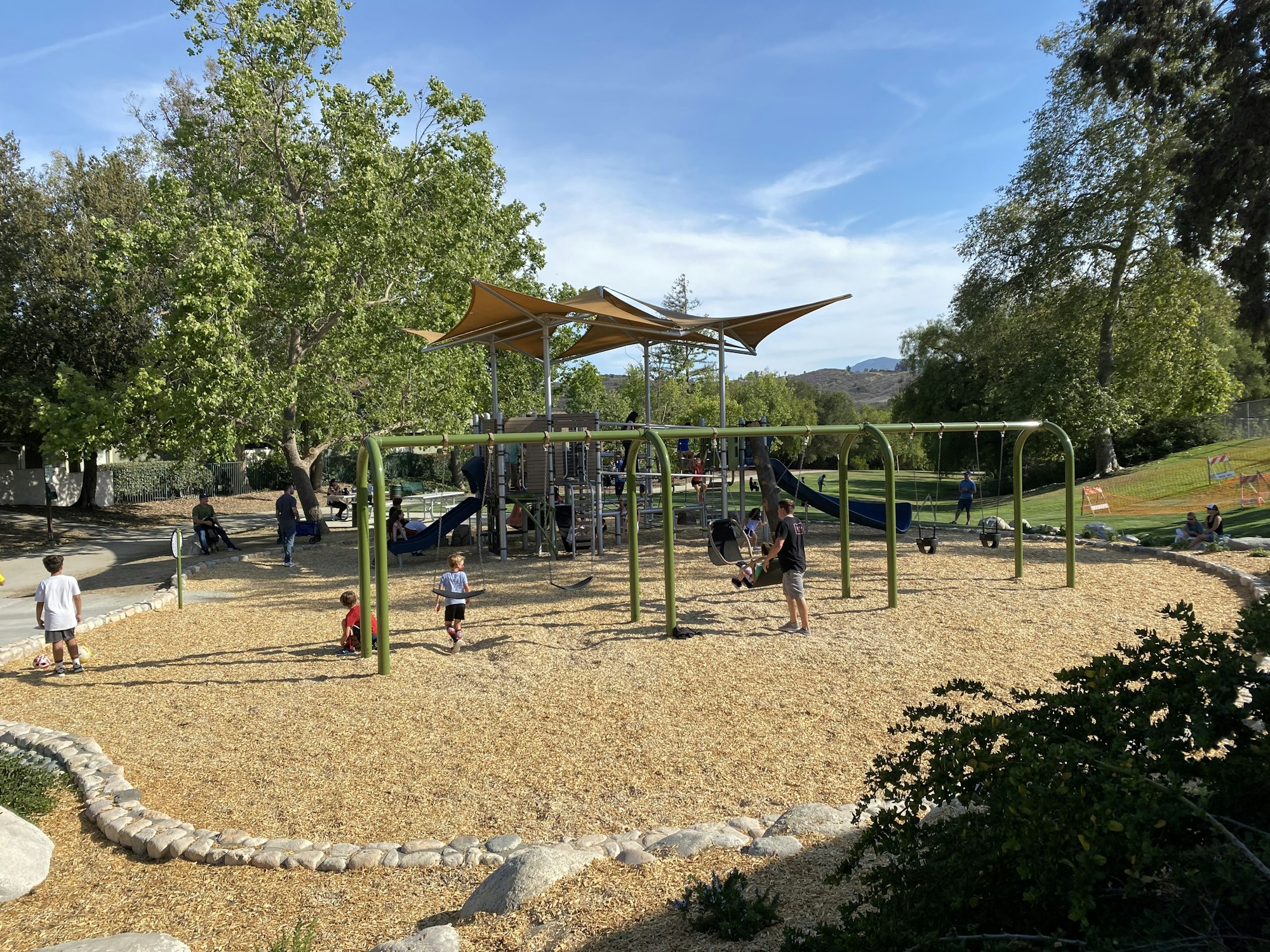 A playground with children playing, swings, and a slide. Trees and a clear sky in the background.