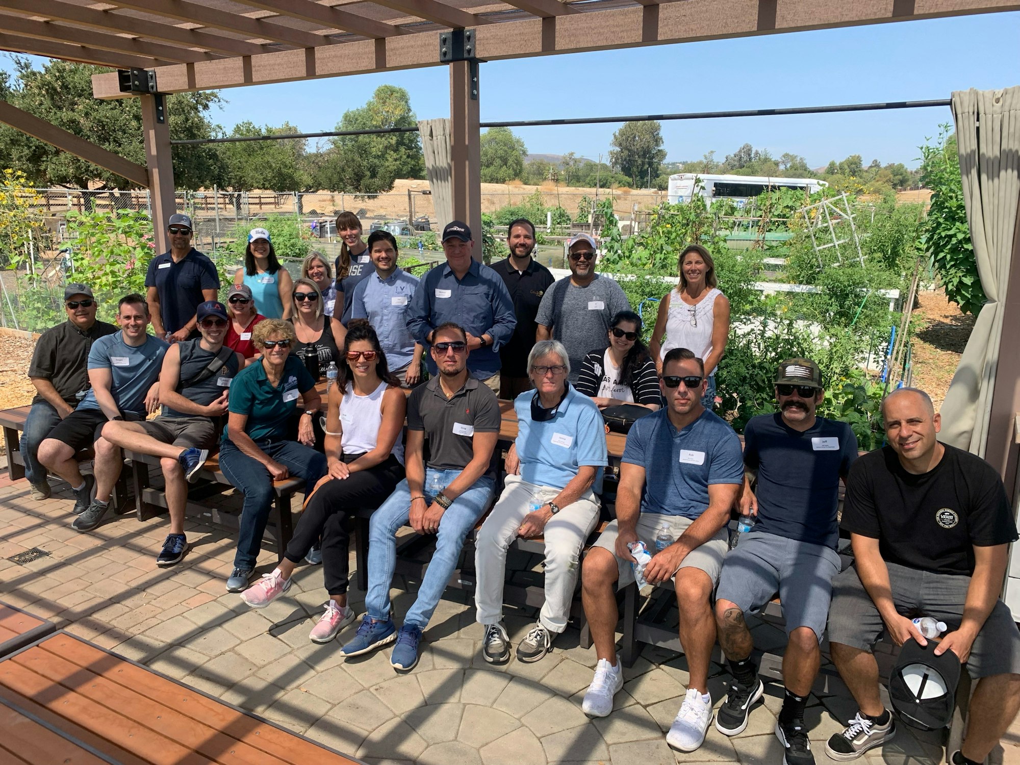 A group of people posed outdoors under a pergola, with plants and trees in the background.