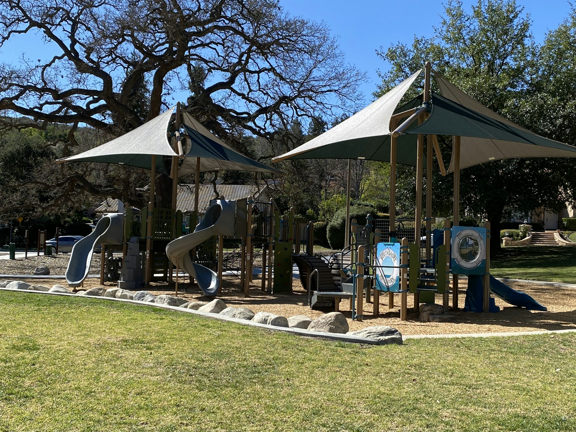 Playground with slides, climbing structures, and shade canopies surrounded by grass and trees.