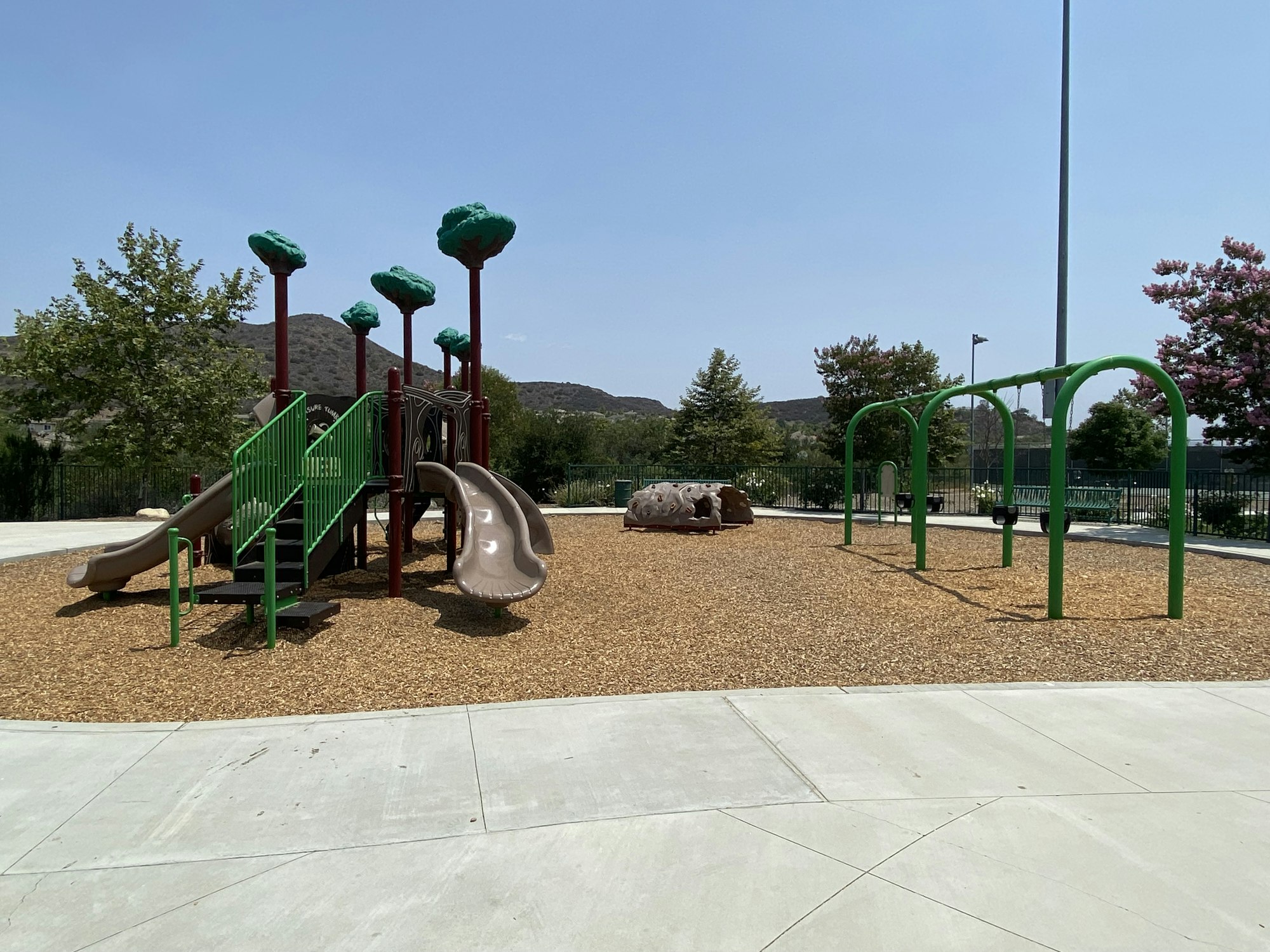 Playground with slides, swings, and climbing structures on a mulch surface, surrounded by greenery and hills.
