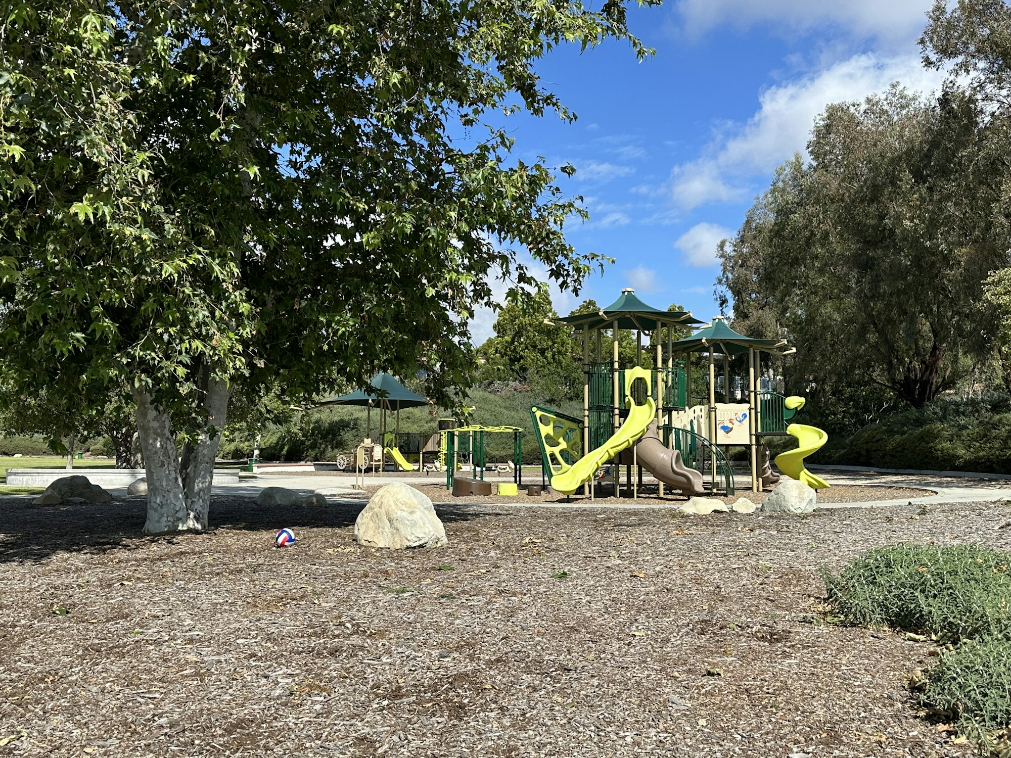A playground with slides and climbing structures, surrounded by trees and rocks, set in a sunny outdoor environment.