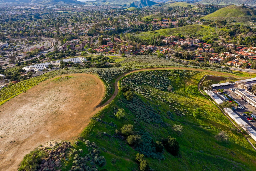 An aerial view of green hills, residential areas, and a parking lot, showcasing nature and urban development.