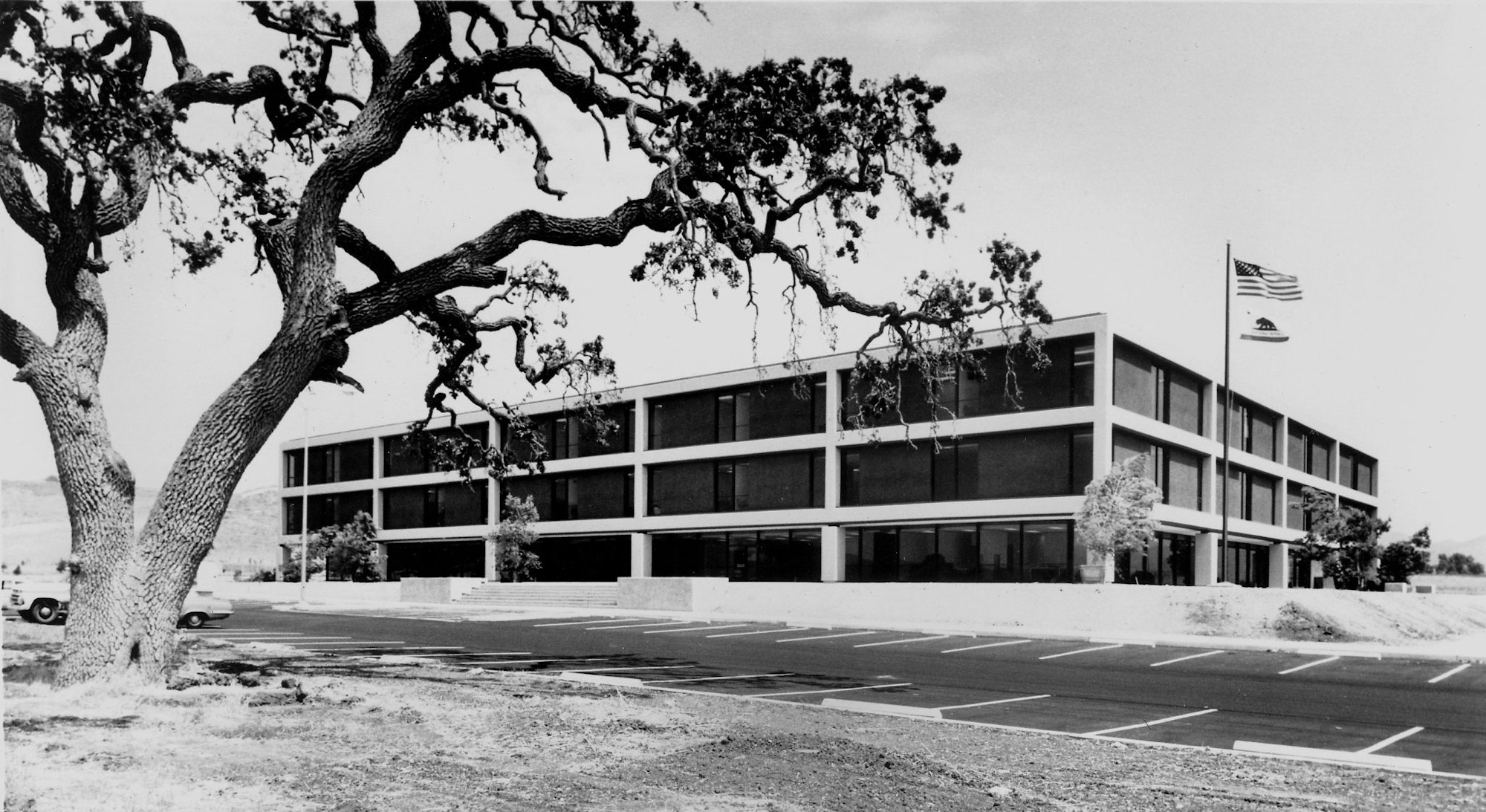 A large, three-story office building with two flags flying in front, surrounded by trees, and an empty parking lot nearby.