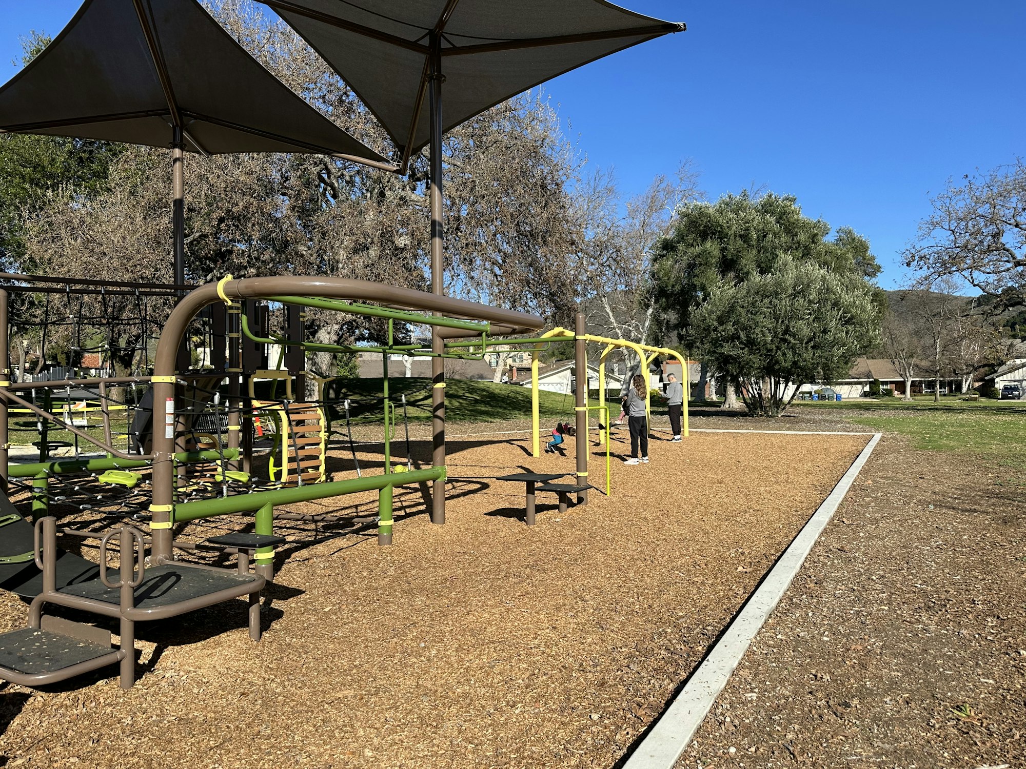 Playground with climbing structures, shade canopies, and people in a park setting under a clear blue sky.